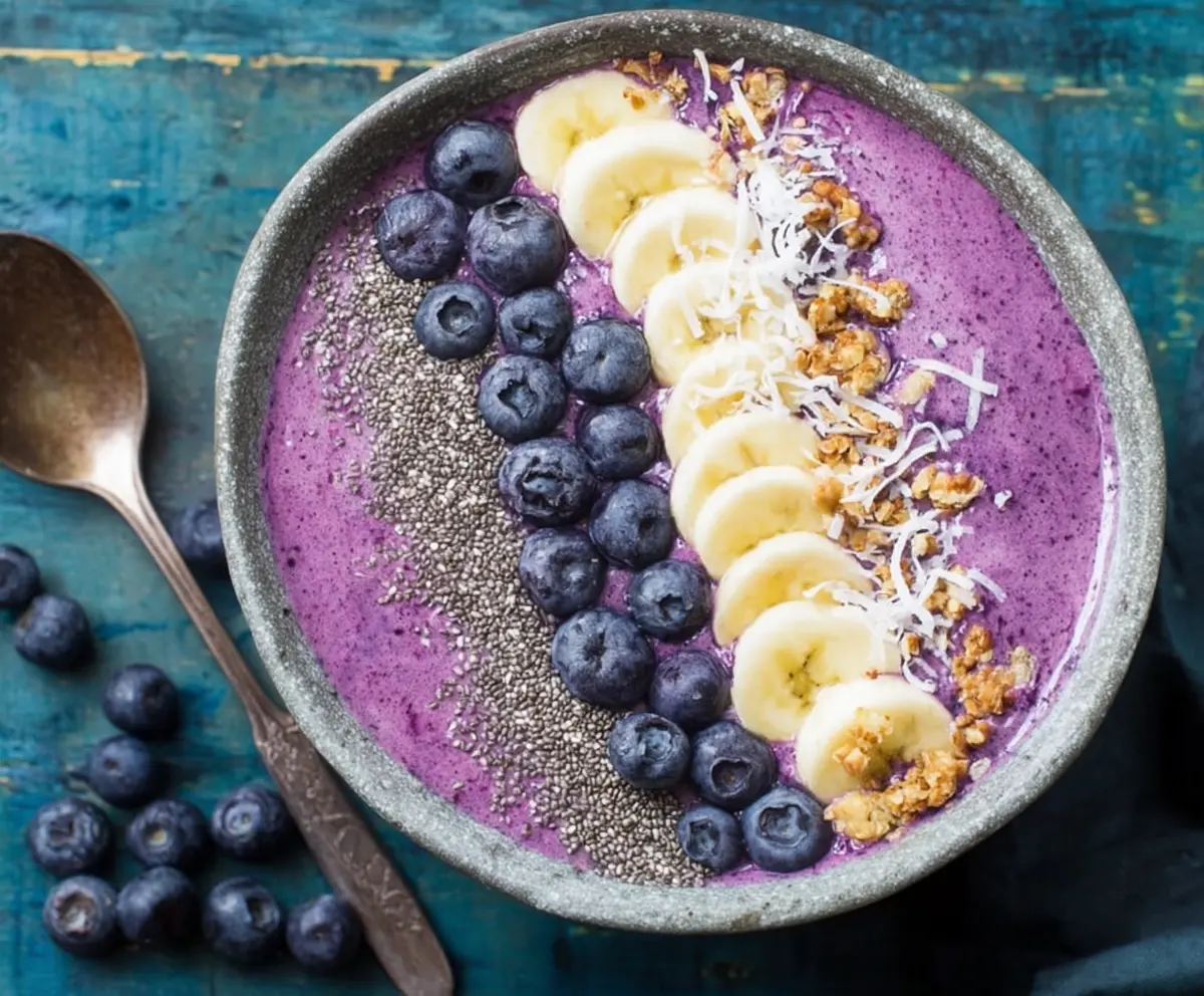 Colorful blueberry smoothie bowl topped with fresh blueberries and granola on a white plate.